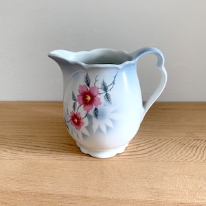 Puede incluir: Una jarra de crema de cerámica blanca con un diseño floral azul y rosa. La jarra tiene un cuerpo redondeado y un asa.