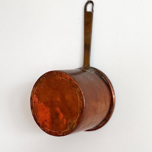 Könnte beinhalten: Ein Kupferkochtopf mit langem Griff und kleiner Aufhängeschlaufe. Der Topf hat einen abgerundeten Boden und einen zylindrischen Körper. Das Kupfer hat eine warme, rotbraune Farbe und weist Altersspuren auf.