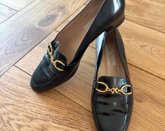 May include: Pair of black patent leather loafers with gold-tone chain detail. The shoes have a low heel and a classic design. The loafers are displayed on a wooden surface.