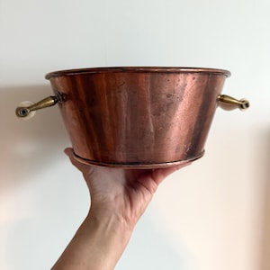 Peut inclure: Seau en métal de couleur cuivre avec un bord arrondi et un corps évasé. Le seau a deux petites poignées avec des accents en laiton et blancs. Le seau est tenu par une main.