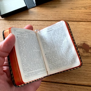 May include: A miniature book, open to reveal printed text, held in a hand. The book features red page edges and a black cover with gold trim. A black box is visible in the background, set on a wooden surface.