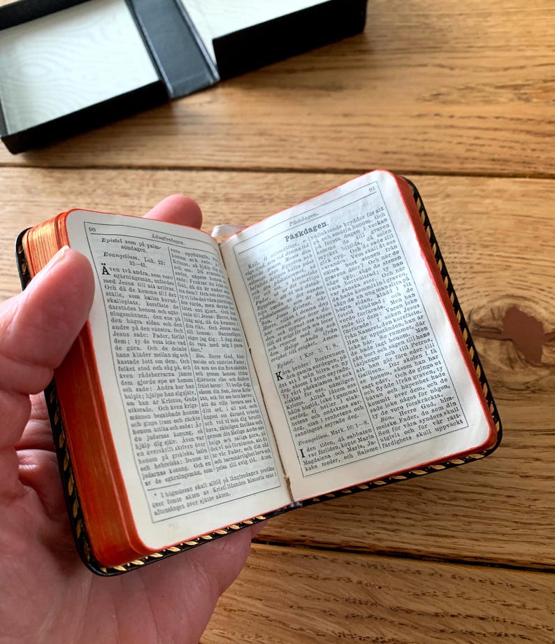 May include: A miniature book, open to reveal printed text, held in a hand. The book features red page edges and a black cover with gold trim. A black box is visible in the background, set on a wooden surface.