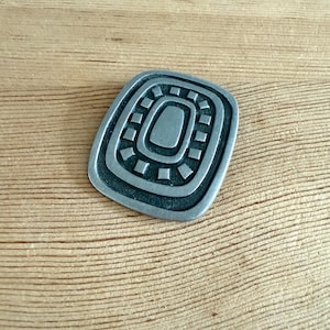 May include: A silver-toned, rectangular brooch with a geometric design. The brooch features a central oval shape surrounded by concentric rectangles and square accents. The metal has a slightly aged appearance, giving it a vintage feel.