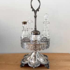 May include: A silver-plated condiment cruet set featuring a central carrying handle, a circular filigree frame, and a scalloped base. The set includes three clear glass bottles with stoppers and a central glass jar with a metal lid.