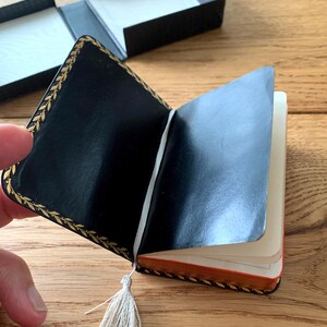 May include: Open black leather-bound notebook with gold leaf trim and a cream-coloured tassel. The notebook is partially open, revealing cream-coloured pages with orange edges. A black box is in the background.