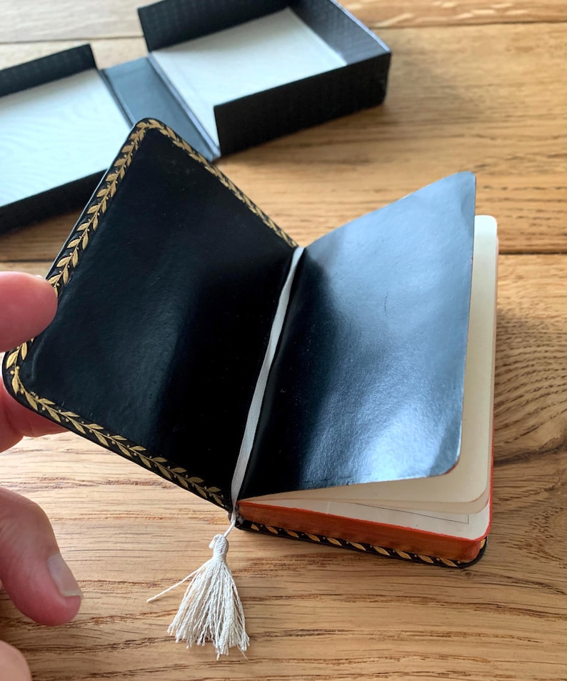 May include: Open black leather-bound notebook with gold leaf trim and a cream-coloured tassel. The notebook is partially open, revealing cream-coloured pages with orange edges. A black box is in the background.