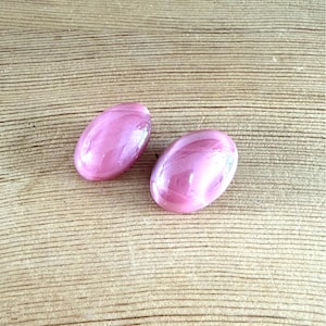 Op de afbeelding: Twee ovale, roze cabochonstenen met een glanzende afwerking. Ze liggen op een lichtbruine houten achtergrond. De stenen hebben een parelmoer effect en reflecteren licht. Elke steen is ongeveer 2 cm lang.