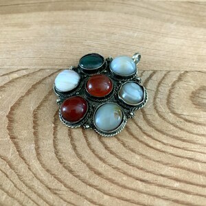 Puede incluir: Un colgante plateado con siete piedras preciosas pulidas de varios colores. Las piedras incluyen tonos de rojo, verde y blanco, engastadas en un marco decorativo calado. El colgante tiene un lazo para una cadena.