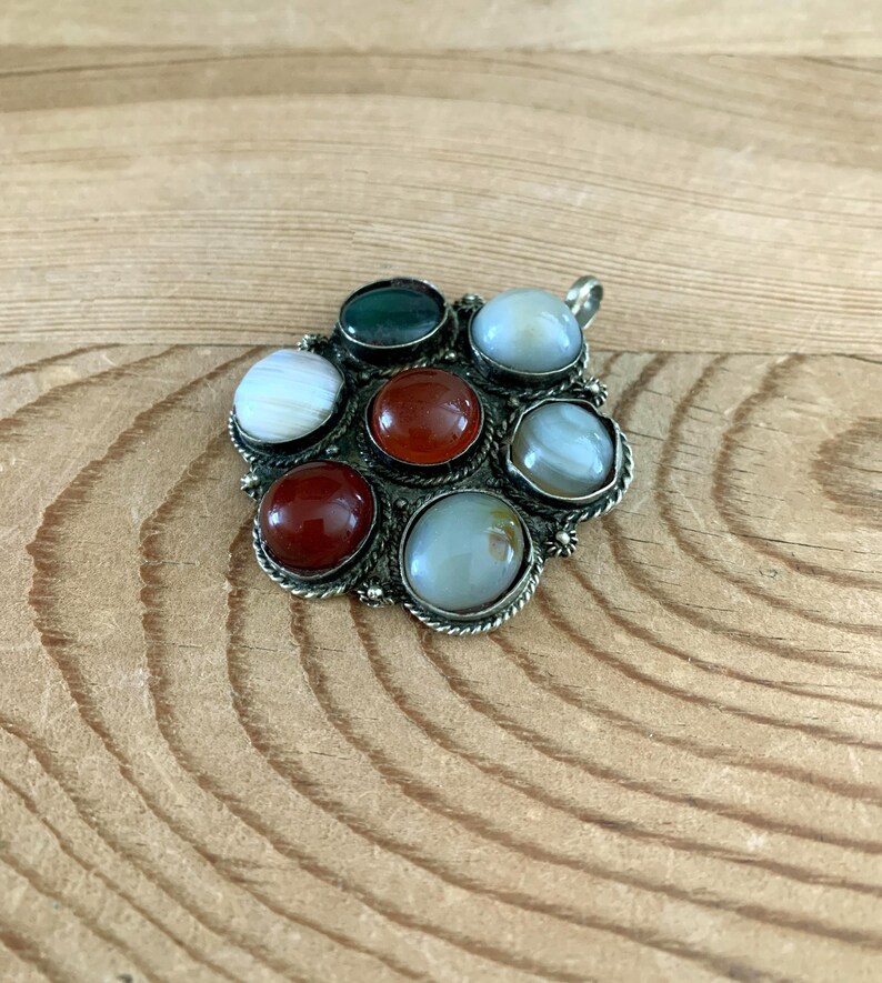Puede incluir: Un colgante plateado con siete piedras preciosas pulidas de varios colores. Las piedras incluyen tonos de rojo, verde y blanco, engastadas en un marco decorativo calado. El colgante tiene un lazo para una cadena.