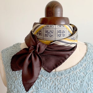 May include: A brown silk scarf with a geometric pattern in shades of yellow, gray, and white. The scarf is tied in a bow around a mannequin's neck.