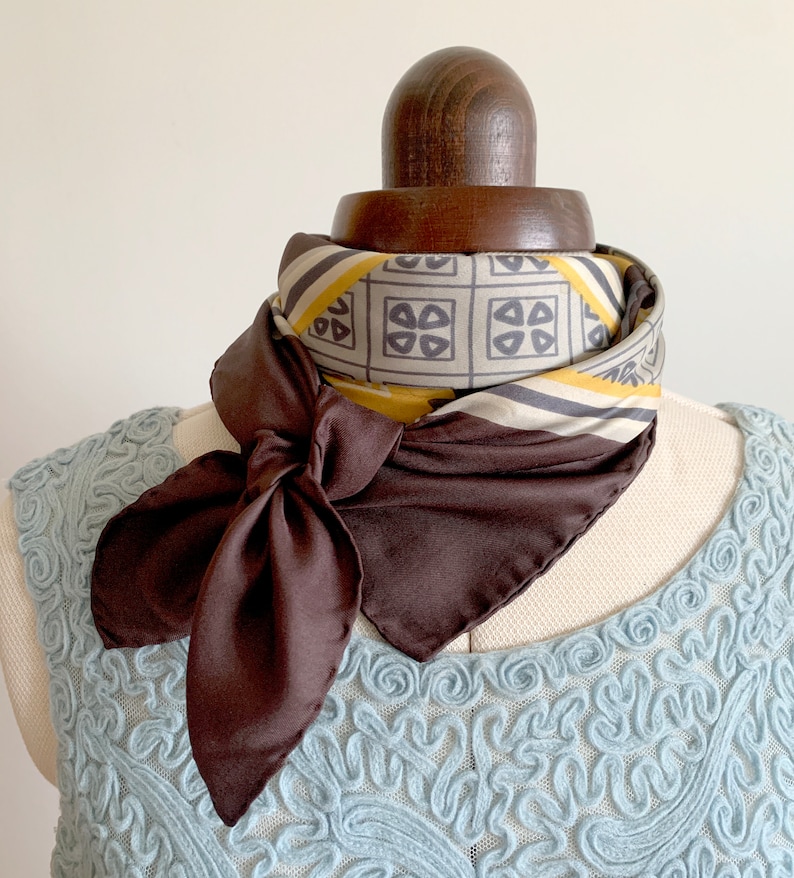 May include: A brown silk scarf with a geometric pattern in shades of yellow, gray, and white. The scarf is tied in a bow around a mannequin's neck.