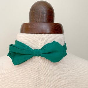 May include: A dark green bow tie displayed on a mannequin. The fabric bow tie is tied in a classic bow shape. The mannequin has a wooden neck and head.