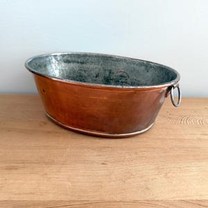 Może przedstawiać: Owalna misa z polerowanym miedzianym wykończeniem na zewnątrz i patynowanym, metalicznym wnętrzem. Naczynie posiada mały, okrągły metalowy uchwyt w kształcie pierścienia przymocowany z jednej strony, stanowiący funkcjonalny detal miedzianego korpusu.