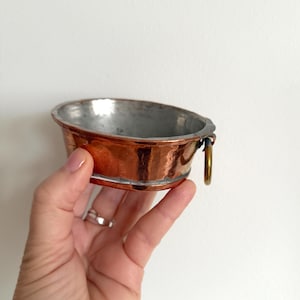 May include: A small, hammered copper bowl with a silver interior. The bowl has a rounded shape with a small handle and a gold-colored ring. The copper has a rich, reddish-brown color, and the bowl appears to be vintage.