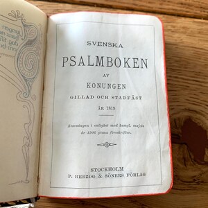 May include: An open book titled "SVENSKA PSALMBOKEN" in black text on a white page. The book has a red spine and is from 1819, published in Stockholm. The page has decorative elements.