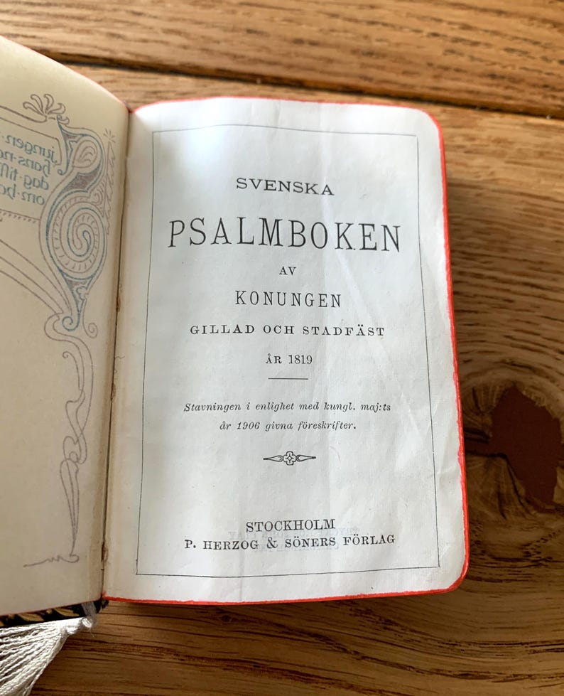 May include: An open book titled "SVENSKA PSALMBOKEN" in black text on a white page. The book has a red spine and is from 1819, published in Stockholm. The page has decorative elements.