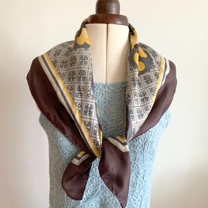 May include: A brown silk scarf with a geometric pattern in yellow and gray. The scarf is tied around a mannequin's neck.