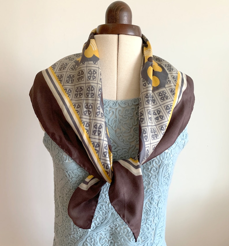 May include: A brown silk scarf with a geometric pattern in yellow and gray. The scarf is tied around a mannequin's neck.