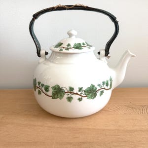 May include: White ceramic teapot with a black handle and a lid. The teapot is decorated with green ivy leaves and vines. The handle is wrapped with a brown material. The teapot is sitting on a wooden surface.
