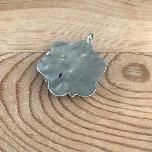 Puede incluir: Un colgante plateado con forma de flor. El colgante tiene una superficie texturizada y un peque&ntilde;o lazo para una cadena. Los bordes est&aacute;n detallados con peque&ntilde;os elementos decorativos. El colgante se muestra sobre una superficie de madera.