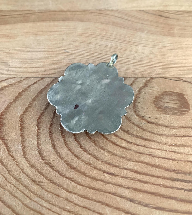 Puede incluir: Un colgante plateado con forma de flor. El colgante tiene una superficie texturizada y un peque&ntilde;o lazo para una cadena. Los bordes est&aacute;n detallados con peque&ntilde;os elementos decorativos. El colgante se muestra sobre una superficie de madera.