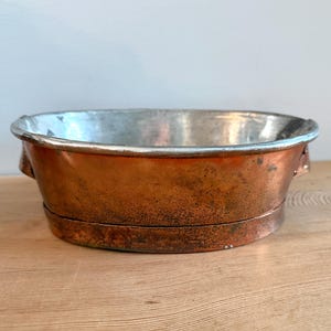 Op de afbeelding: Ovale koperen waskom met een zilveren binnenkant. De waskom heeft twee kleine handvatten aan de zijkanten en een iets verhoogde bodem. Het koper heeft een rijke, verouderde patina en de binnenkant is glad en reflecterend. De waskom staat op een houten oppervlak.