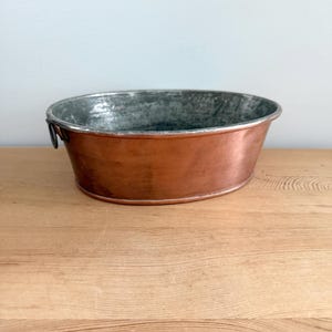 Puede incluir: Un recipiente de forma ovalada con un exterior de cobre pulido y un interior de tono plateado metálico desgastado. El cuenco incluye una pequeña asa circular oscura unida al lateral, mostrando un contraste entre el acabado de cobre cálido y la superficie interior fría y texturizada.