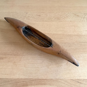 Op de afbeelding: Een vintage houten weefspoel met een puntig uiteinde en een centrale holte voor het vasthouden van de inslagdraad. De spoel heeft een warme bruine kleur en vertoont tekenen van slijtage, wat duidt op gebruik in de textielproductie. De spoel is ongeveer 25 cm lang.