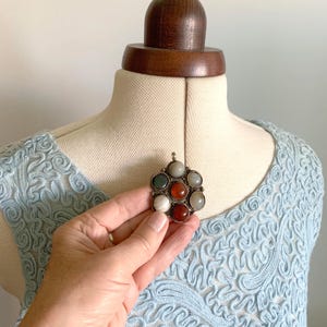 Puede incluir: Un colgante plateado con un dise&ntilde;o floral, con siete piedras preciosas de colores en tonos rojos, verdes y blancos. El colgante se muestra sobre un top de encaje azul claro, resaltando los detalles intrincados y los colores vibrantes.