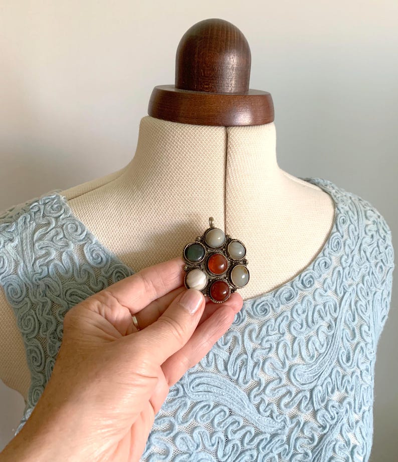 Puede incluir: Un colgante plateado con un dise&ntilde;o floral, con siete piedras preciosas de colores en tonos rojos, verdes y blancos. El colgante se muestra sobre un top de encaje azul claro, resaltando los detalles intrincados y los colores vibrantes.