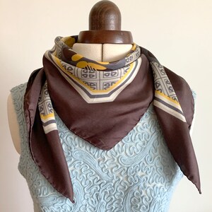 May include: A brown silk scarf with a geometric pattern in yellow, gray, and white. The scarf is tied around a mannequin's neck.
