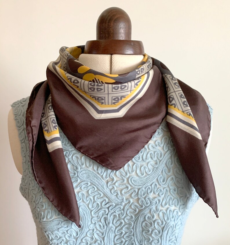 May include: A brown silk scarf with a geometric pattern in yellow, gray, and white. The scarf is tied around a mannequin's neck.