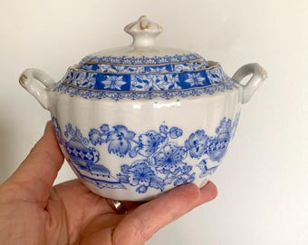 May include: A white porcelain sugar bowl with a blue and gold floral design. The bowl has a lid and two handles.