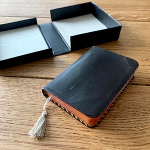 May include: A black leather-bound book with a tassel bookmark, next to an open black box. The book has a worn appearance, with a light-coloured tassel. The box is open, revealing a white interior. The items are on a wooden surface.