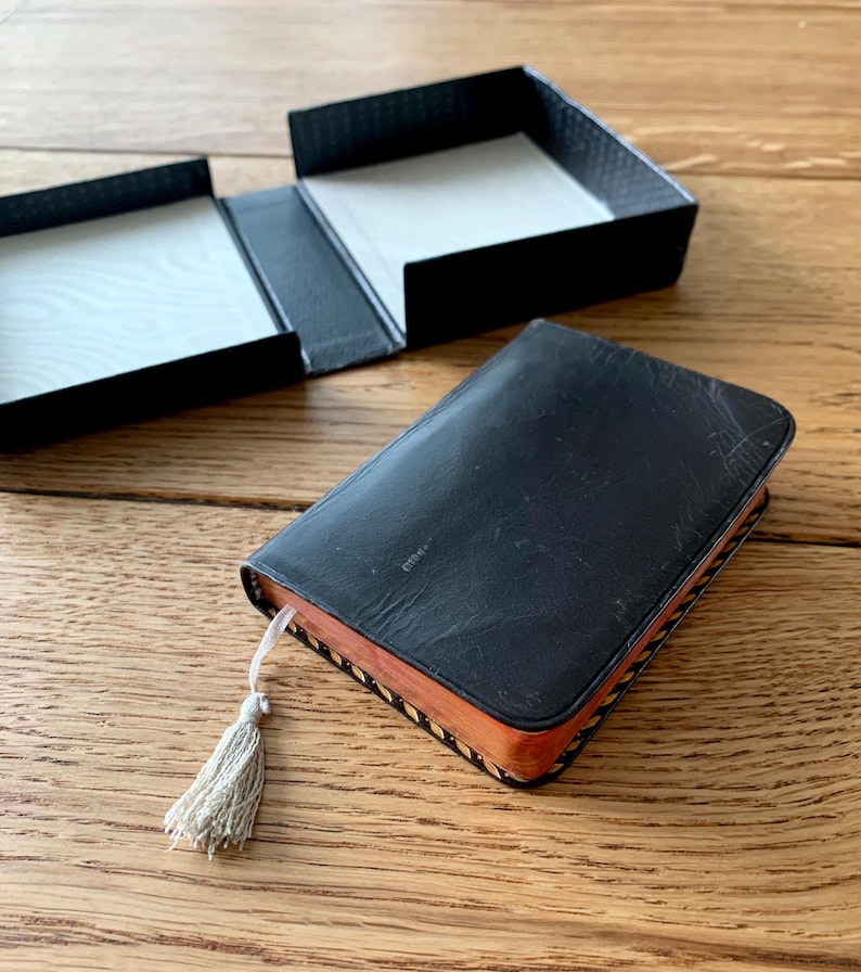 May include: A black leather-bound book with a tassel bookmark, next to an open black box. The book has a worn appearance, with a light-coloured tassel. The box is open, revealing a white interior. The items are on a wooden surface.