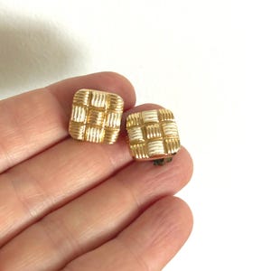 Puede incluir: Un par de pendientes cuadrados dorados con diseño tejido. Los pendientes tienen una superficie texturizada con secciones doradas y blancas alternas. Los cierres de clip son visibles. Estos pendientes vintage son un accesorio clásico.