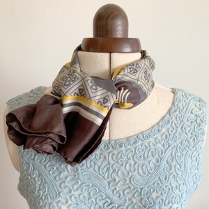 May include: A brown and gray patterned silk scarf with a yellow stripe. The scarf is tied around a dress form's neck and the ends are draped down the front of the form.