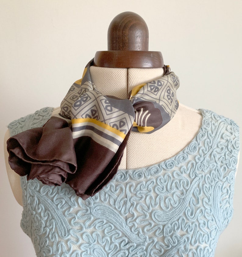 May include: A brown and gray patterned silk scarf with a yellow stripe. The scarf is tied around a dress form's neck and the ends are draped down the front of the form.