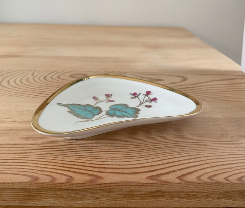 May include: A white ceramic trinket dish with a gold rim and a floral design featuring teal leaves and pink flowers.