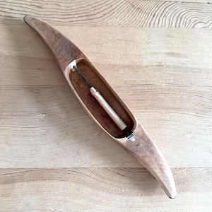 Op de afbeelding: Een houten weefspoel, een hulpmiddel voor het weven. Het heeft een langwerpige, gebogen vorm en is gemaakt van lichtbruin hout. Binnenin bevindt zich een witte spoel die door een zwarte draad wordt vastgehouden.