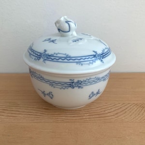 May include: A white porcelain sugar bowl with a lid, adorned with blue floral and wave patterns. The bowl has a rounded shape and a small, decorative handle on the lid. The design is delicate and classic, suitable for serving sugar or other small items.