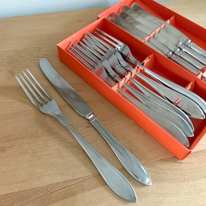 Może przedstawiać: Zestaw sztućców ze stali nierdzewnej, w tym widelce i noże, prezentowany w pomarańczowym pudełku. Sztućce mają prosty, elegancki design z długimi, smukłymi uchwytami. Zestaw jest ułożony na jasnej drewnianej powierzchni.