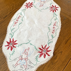 May include: White linen table runner with embroidered red poinsettia flowers, green vines, and two figures of Santa Claus. The text "God Jul" is embroidered in red on each side of the runner.