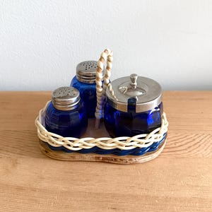 Puede incluir: Un juego vintage de salero y pimentero azul cobalto y un azucarero con tapa plateada, presentado en una bandeja tejida con asa. El juego está diseñado para uso en la mesa y añade un toque retro a cualquier comedor.