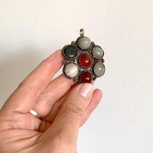 Puede incluir: Colgante plateado con dise&ntilde;o floral, con ocho piedras preciosas redondas y coloridas. Las piedras preciosas incluyen tonos de rojo, verde, gris y blanco. El colgante se muestra sobre un fondo blanco.