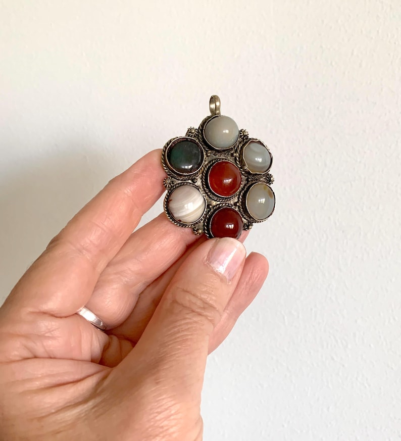 Puede incluir: Colgante plateado con dise&ntilde;o floral, con ocho piedras preciosas redondas y coloridas. Las piedras preciosas incluyen tonos de rojo, verde, gris y blanco. El colgante se muestra sobre un fondo blanco.