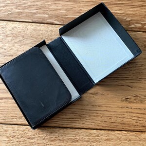 May include: A black rectangular box with a hinged lid, open to reveal a white interior. A black wallet sits inside the box. The wallet and box appear to be made of leather.
