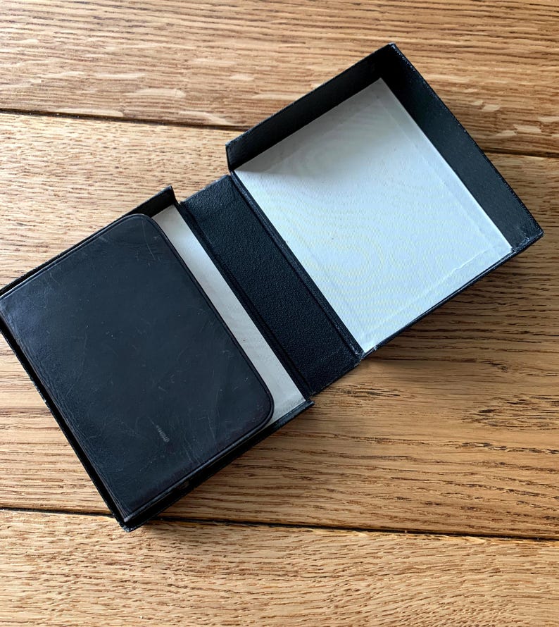 May include: A black rectangular box with a hinged lid, open to reveal a white interior. A black wallet sits inside the box. The wallet and box appear to be made of leather.