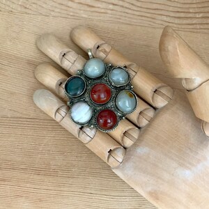 Puede incluir: Un colgante de plata con un dise&ntilde;o circular que presenta siete piedras preciosas de colores. Las piedras preciosas incluyen piedras rojas, verdes, blancas y azul claro. El colgante se muestra en una mano de madera.