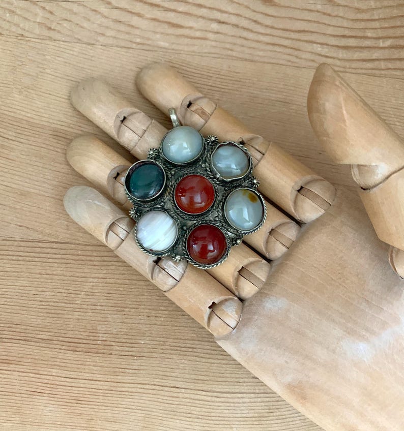 Puede incluir: Un colgante de plata con un dise&ntilde;o circular que presenta siete piedras preciosas de colores. Las piedras preciosas incluyen piedras rojas, verdes, blancas y azul claro. El colgante se muestra en una mano de madera.
